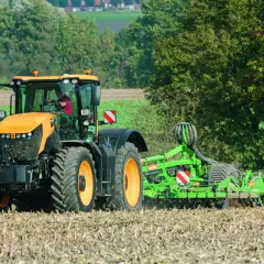 Pomarańczowy JCB Fastrac, pracując z broną, przemierza pole z prędkością 90 km/h, przygotowując ziemię do siewu.