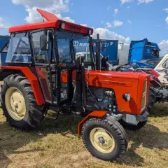 Nowy Ursus C-360 w jaskrawym pomarańczowym kolorze, z kabiną i błyszczącymi felgami, prezentuje się dumnie na tle innych maszyn.