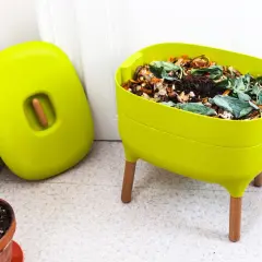 Zielony kompostownik na balkonie, pełen resztek organicznych, obok stoi jego pokrywa.