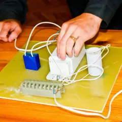 Ręce montują elementy elektryczne, przygotowując się do tego, jak podłączyć pastucha. Widać kondensator, dławik i listwę zaciskową.