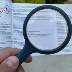 Lupa powiększa etykietę z ostrzeżeniami dotyczącymi środków ochrony roślin. Widoczne są symbole ostrzegawcze i tekst, w tym fraza "piorin komunikaty opryski".