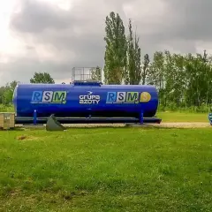 Duży, niebieski zbiornik RSM 32 skład nawozów, obok maszyny rolnicze i drzewa.