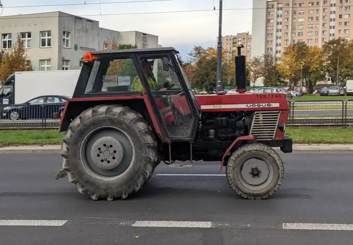 Czerwony Ursus 1954, model 902, jedzie ulicą. W tle budynki mieszkalne i drzewa.