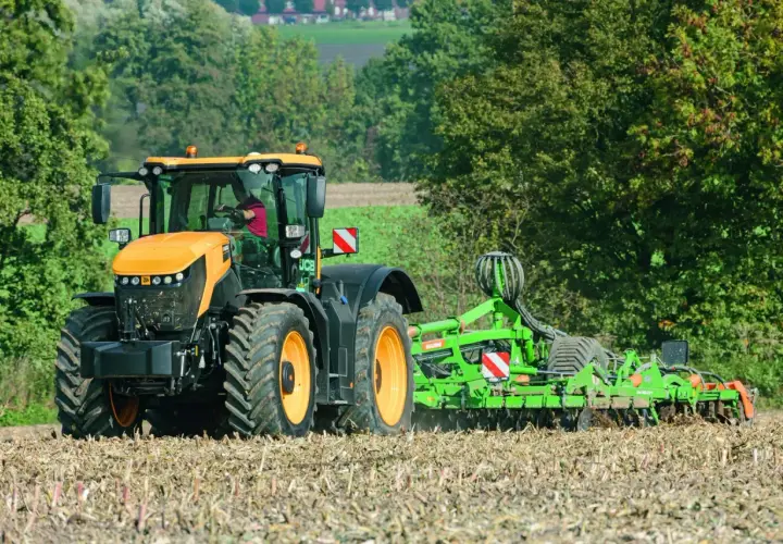 Pomarańczowy JCB Fastrac, pracując z broną, przemierza pole z prędkością 90 km/h, przygotowując ziemię do siewu.