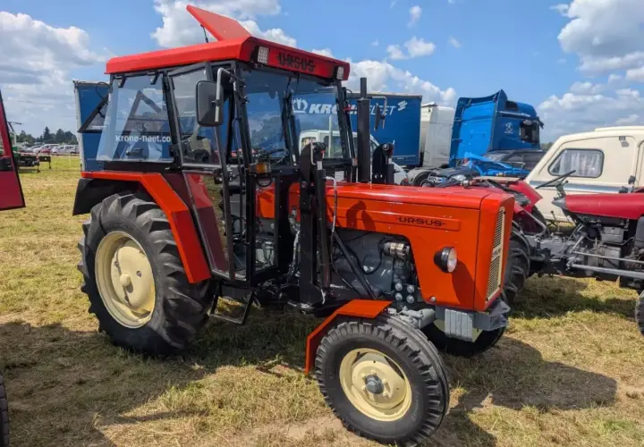 Nowy Ursus C-360 w jaskrawym pomarańczowym kolorze, z kabiną i błyszczącymi felgami, prezentuje się dumnie na tle innych maszyn.