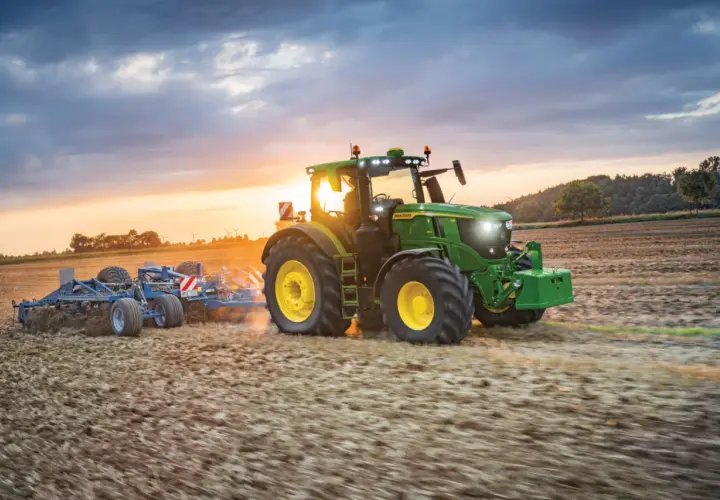 Zielony traktor John Deere Mannheim orze pole o zachodzie słońca.