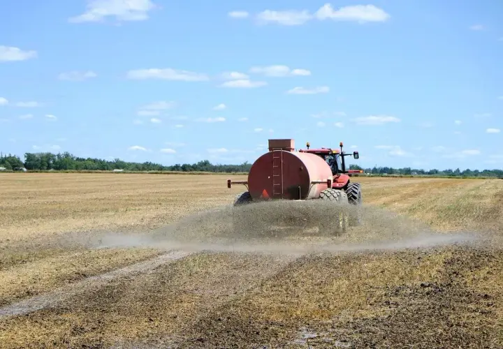 Czerwony traktor z beczkowozem rozprowadza odpad z biogazowni na polu.