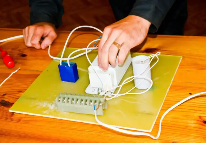 Ręce montują elementy elektryczne, przygotowując się do tego, jak podłączyć pastucha. Widać kondensator, dławik i listwę zaciskową.
