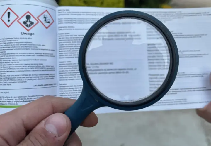 Lupa powiększa etykietę z ostrzeżeniami dotyczącymi środków ochrony roślin. Widoczne są symbole ostrzegawcze i tekst, w tym fraza "piorin komunikaty opryski".