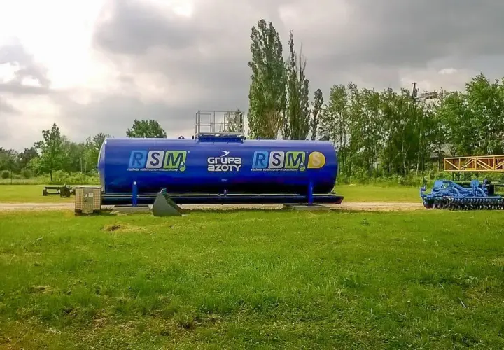 Duży, niebieski zbiornik RSM 32 skład nawozów, obok maszyny rolnicze i drzewa.