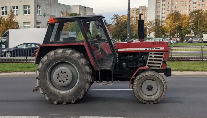 Czerwony Ursus 1954, model 902, jedzie ulicą. W tle budynki mieszkalne i drzewa.