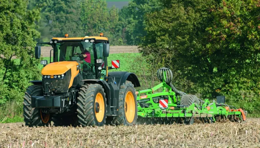 Pomarańczowy JCB Fastrac, pracując z broną, przemierza pole z prędkością 90 km/h, przygotowując ziemię do siewu.