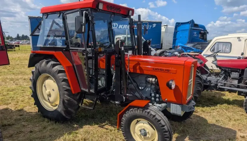 Nowy Ursus C-360 w jaskrawym pomarańczowym kolorze, z kabiną i błyszczącymi felgami, prezentuje się dumnie na tle innych maszyn.