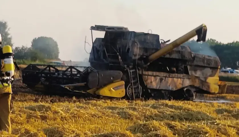Pożar kombajnu na polu. Maszyna jest mocno zniszczona, widać dym i strażaka w akcji.