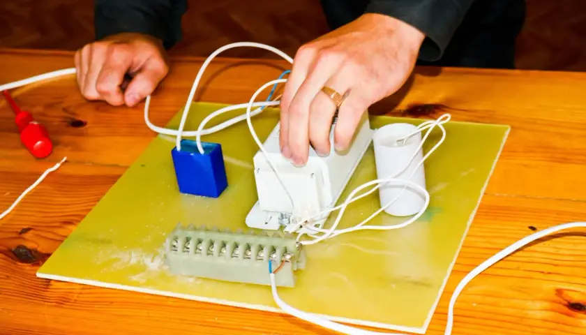 Ręce montują elementy elektryczne, przygotowując się do tego, jak podłączyć pastucha. Widać kondensator, dławik i listwę zaciskową.