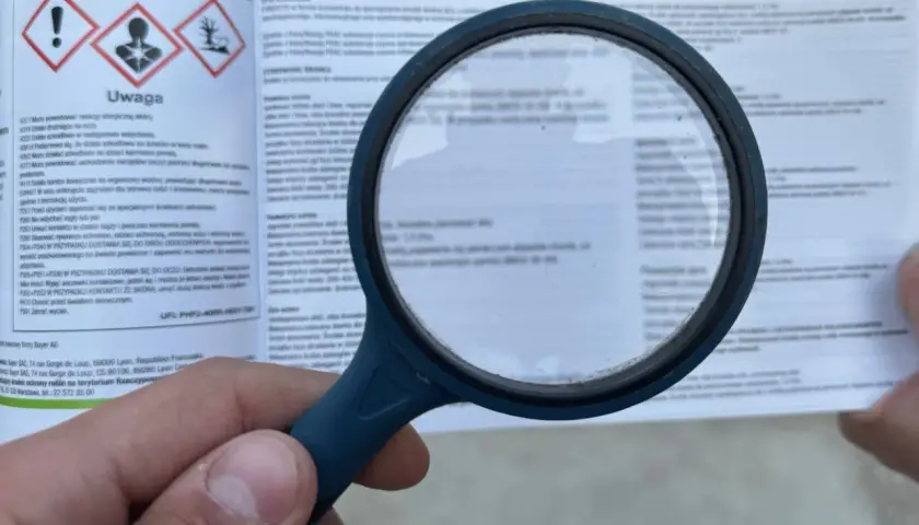 Lupa powiększa etykietę z ostrzeżeniami dotyczącymi środków ochrony roślin. Widoczne są symbole ostrzegawcze i tekst, w tym fraza "piorin komunikaty opryski".