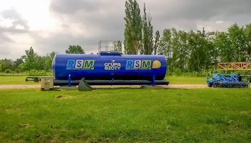 Duży, niebieski zbiornik RSM 32 skład nawozów, obok maszyny rolnicze i drzewa.