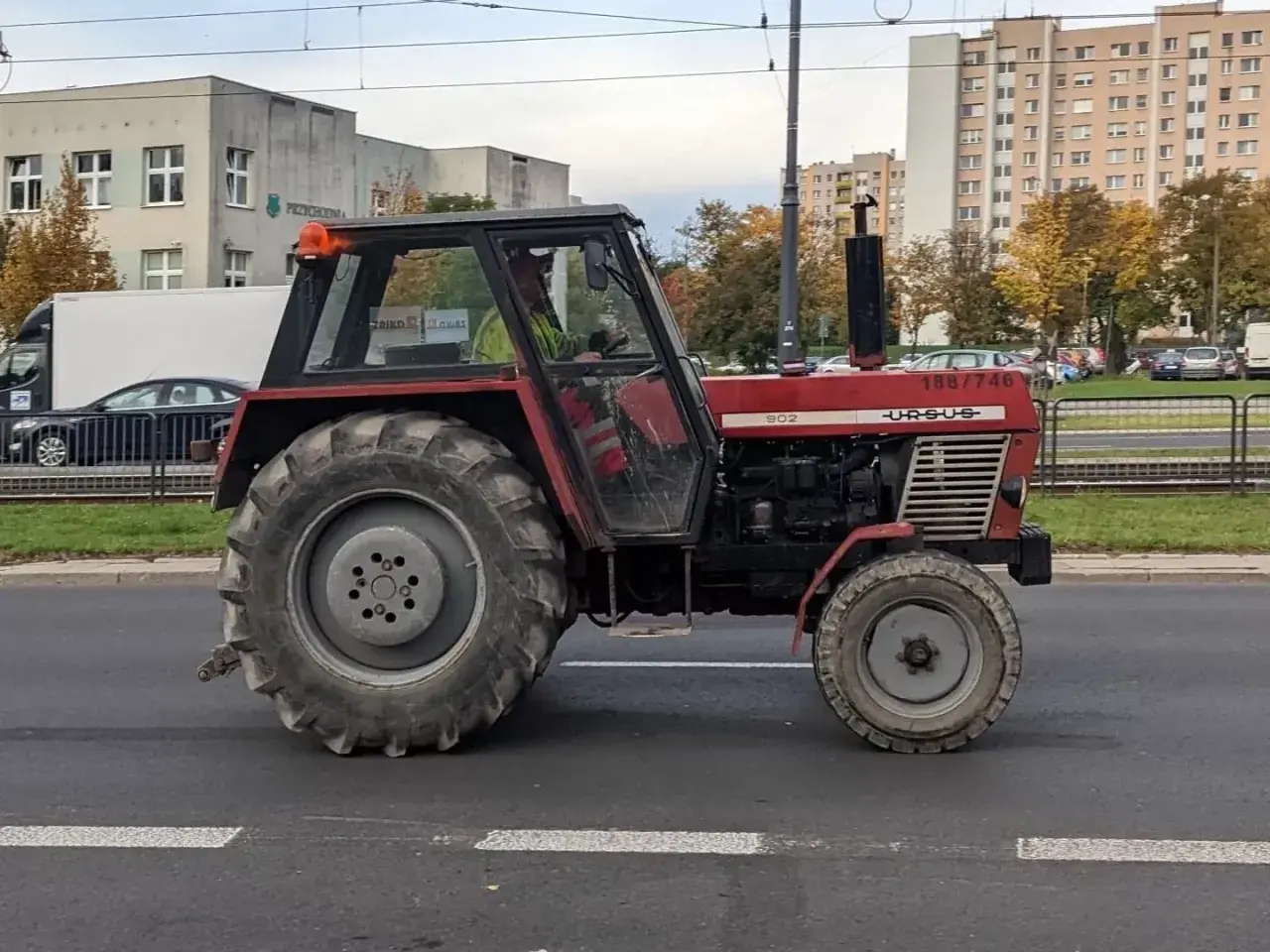 Czerwony Ursus 1954, model 902, jedzie ulicą. W tle budynki mieszkalne i drzewa.