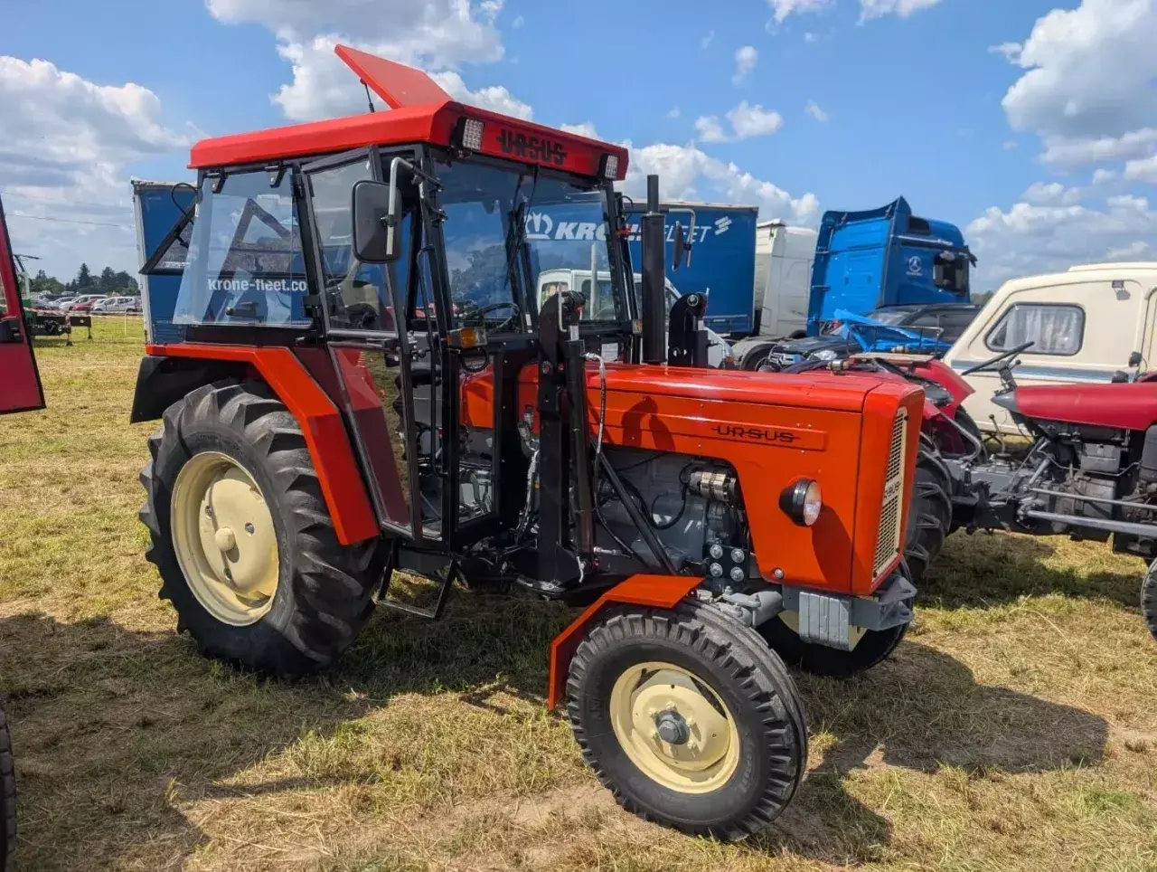 Nowy Ursus C-360 w jaskrawym pomarańczowym kolorze, z kabiną i błyszczącymi felgami, prezentuje się dumnie na tle innych maszyn.