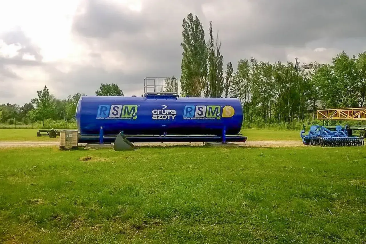 Duży, niebieski zbiornik RSM 32 skład nawozów, obok maszyny rolnicze i drzewa.