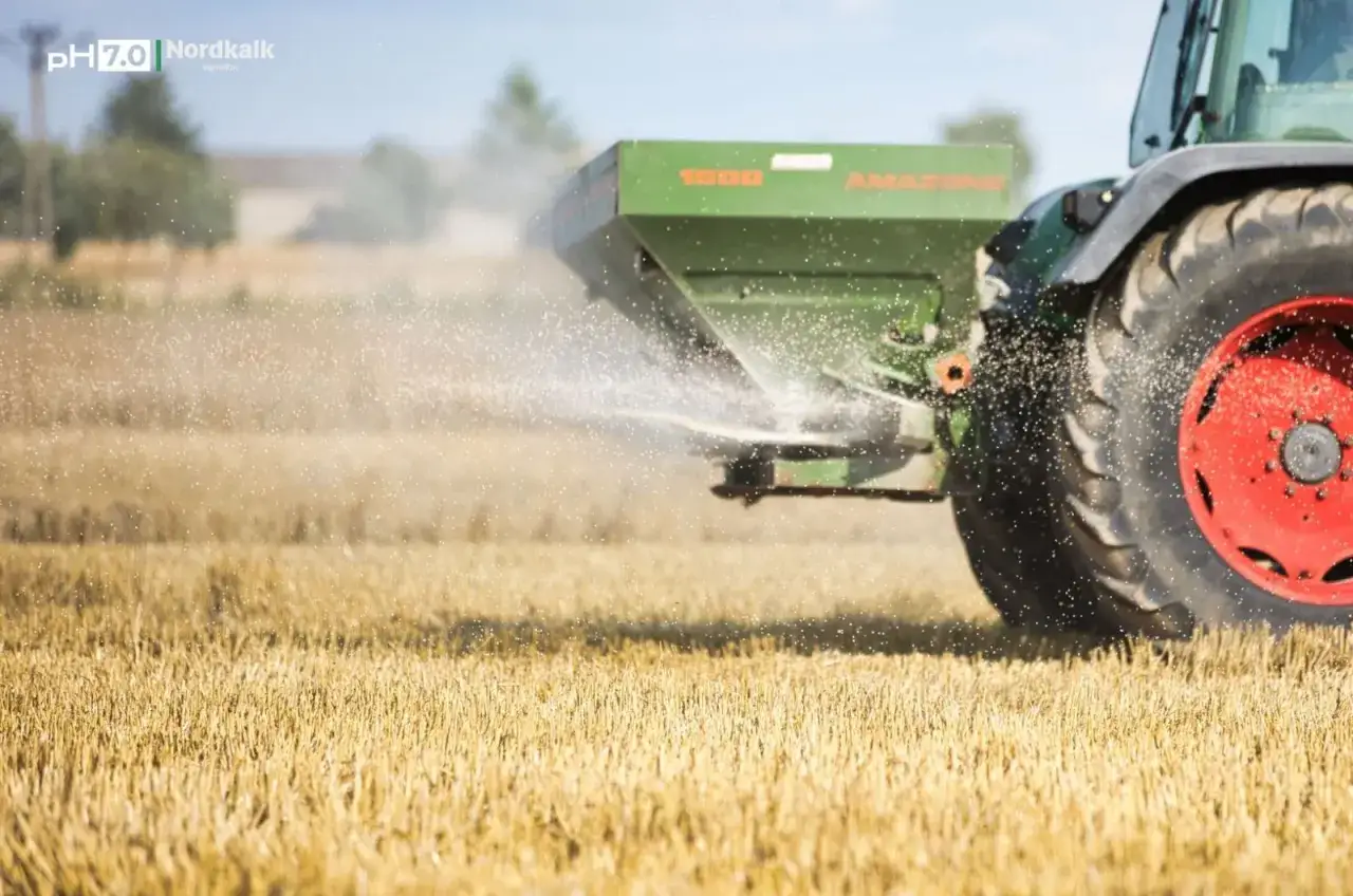 Traktor rozsypuje wapno magnezowe na polu. Drobinki nawozu unoszą się w powietrzu, poprawiając jakość gleby.