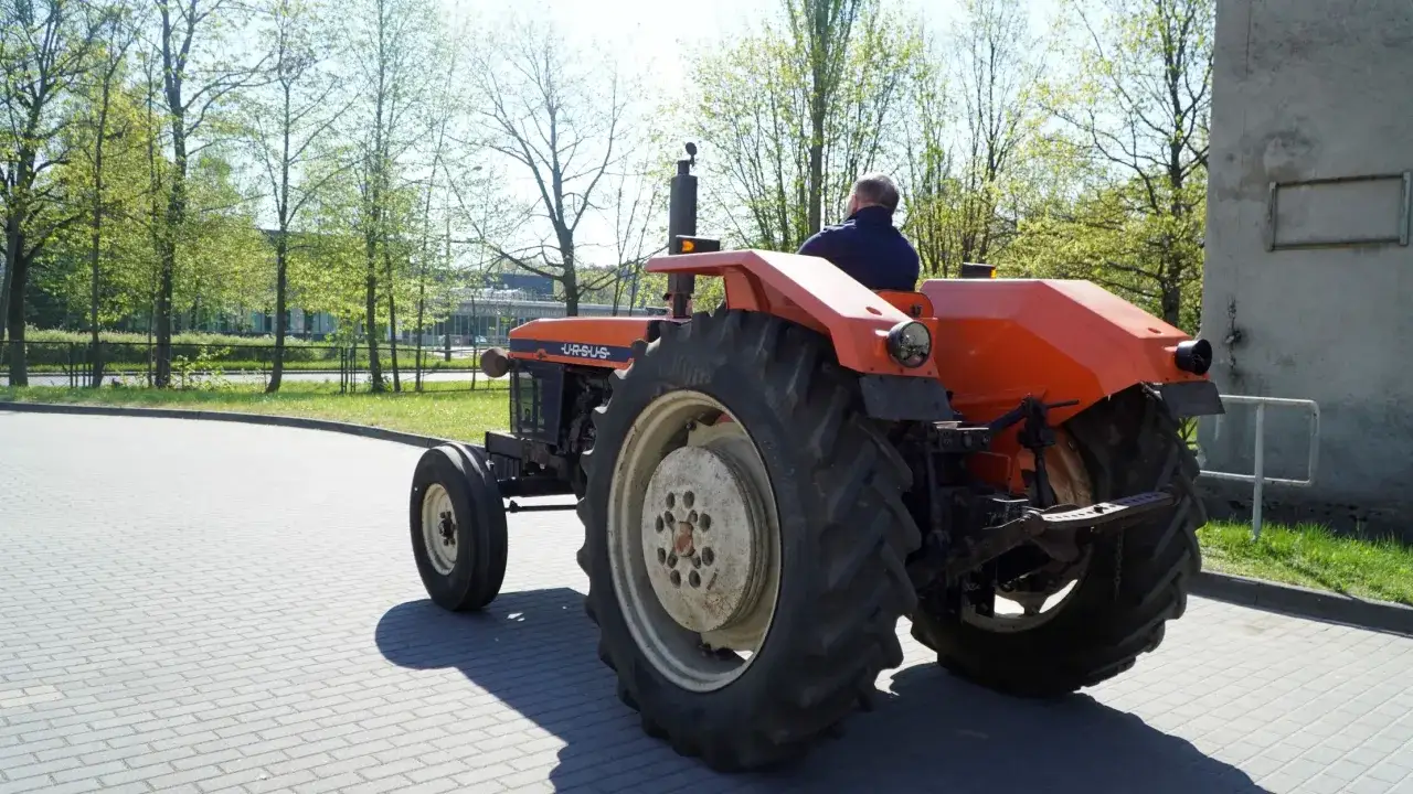 Pomarańczowy traktor Ursus U 710 z kierowcą jedzie po brukowanej drodze, otoczony zielenią i drzewami.
