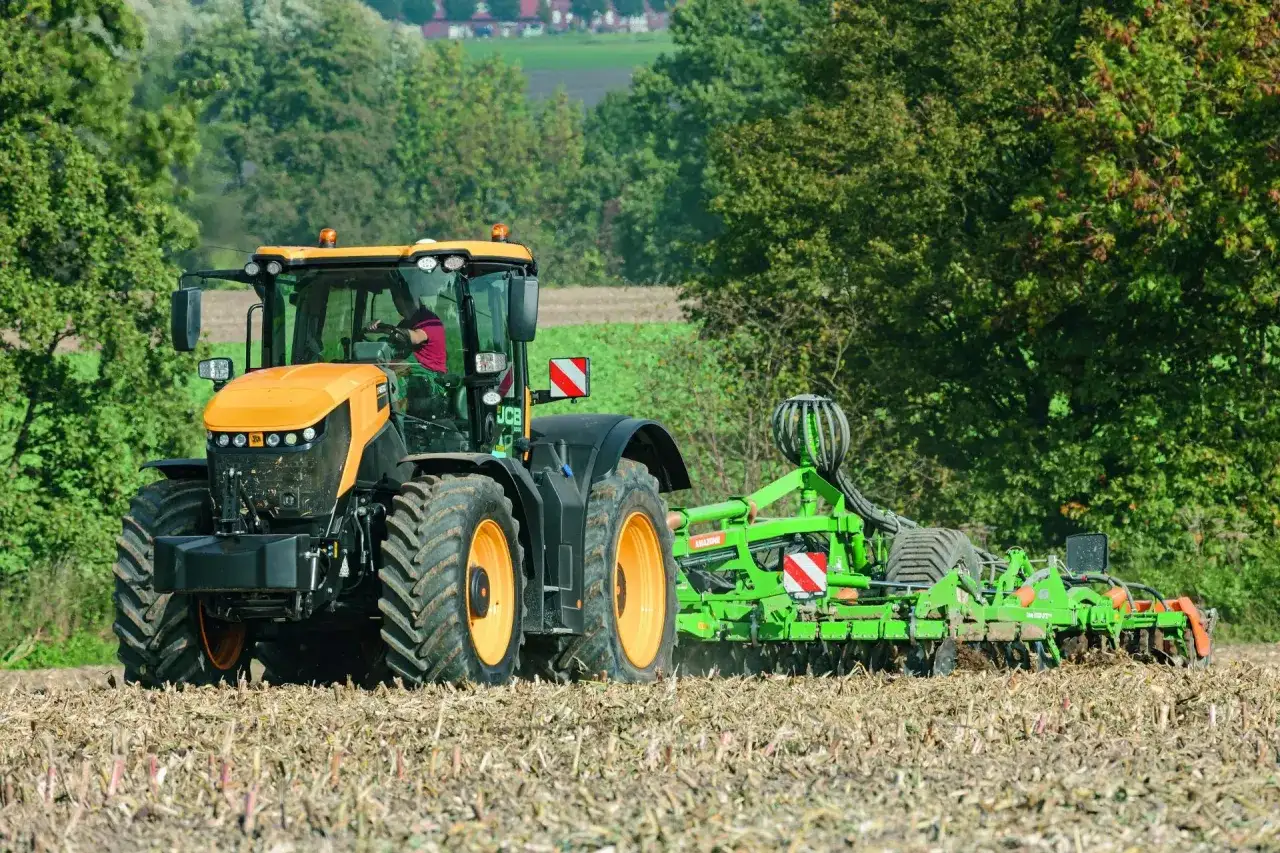 Pomarańczowy JCB Fastrac, pracując z agregatem uprawowym, przemierza pole z prędkością 90 km/h, przygotowując ziemię do siewu.