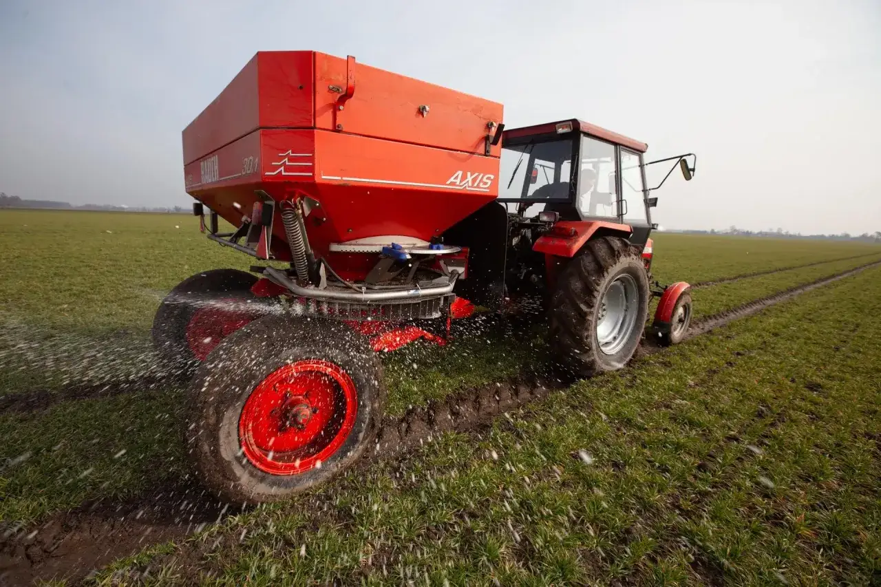 Czerwony traktor z rozsiewaczem nawozu, który rozprowadza mocznik nawóz na zielonym polu.