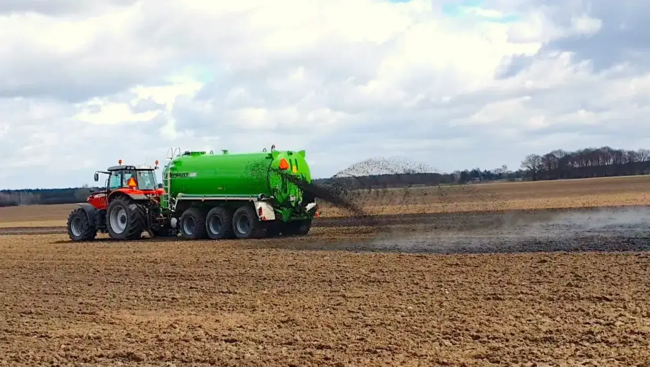 Czerwony traktor z zielonym wozem asenizacyjnym rozprowadza odpad z biogazowni na polu.